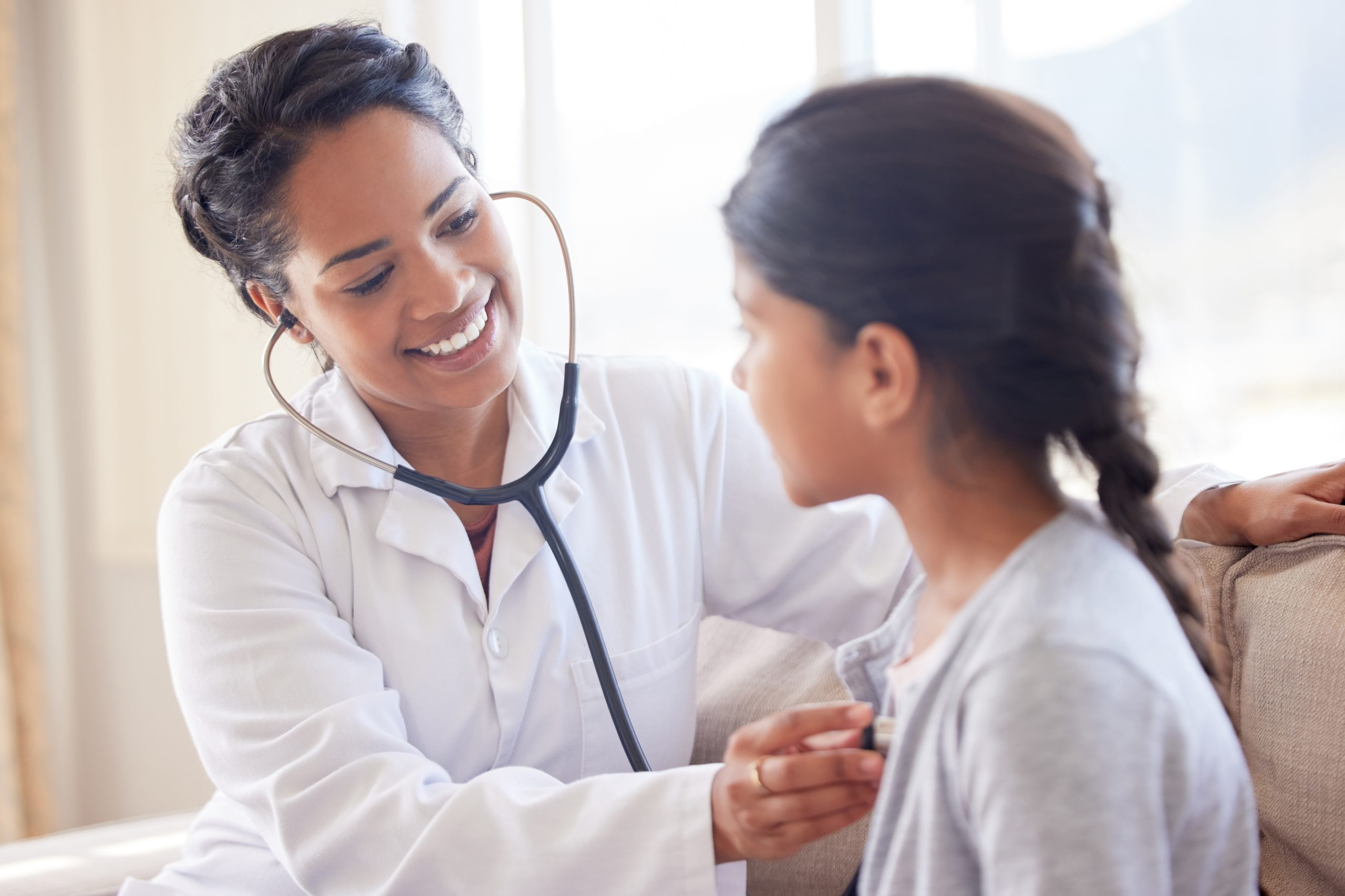 Doctor examining a little girl with stethoscope. Female paediatrician listening to child’s heartbeat during home visit or checkup at clinic. Caring woman in healthcare sector working with patient